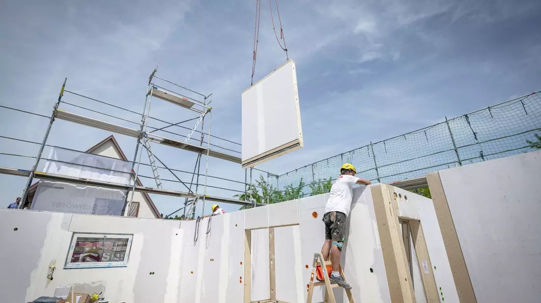 Ein Bauarbeiter steht auf einer Leiter und arbeitet im Rohbau eines Fertighauses, während gerade ein weiteres Wandelement von einem Kran heruntergelassen wird. - Bild: Daniel Maurer/dpa