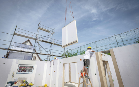 Ein Bauarbeiter steht auf einer Leiter und arbeitet im Rohbau eines Fertighauses, während gerade ein weiteres Wandelement von einem Kran heruntergelassen wird. - Foto: Daniel Maurer/dpa