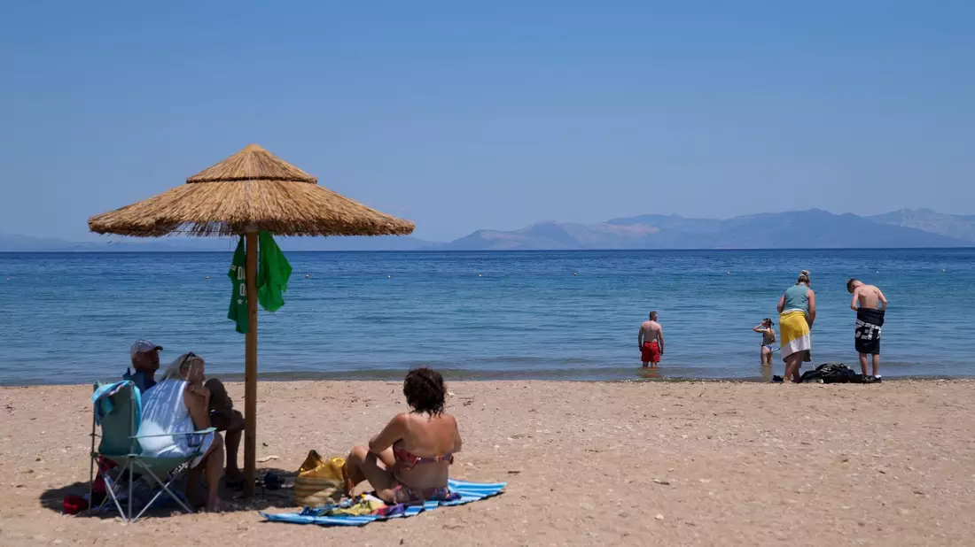Eine Frau mit einem Handventilator unter einem Sonnenschirm am Strand in Griechenland. Die heißen Mittagsstunden sollten möglichst gemieden werden. - Bild: Thanassis Stavrakis/AP/dpa