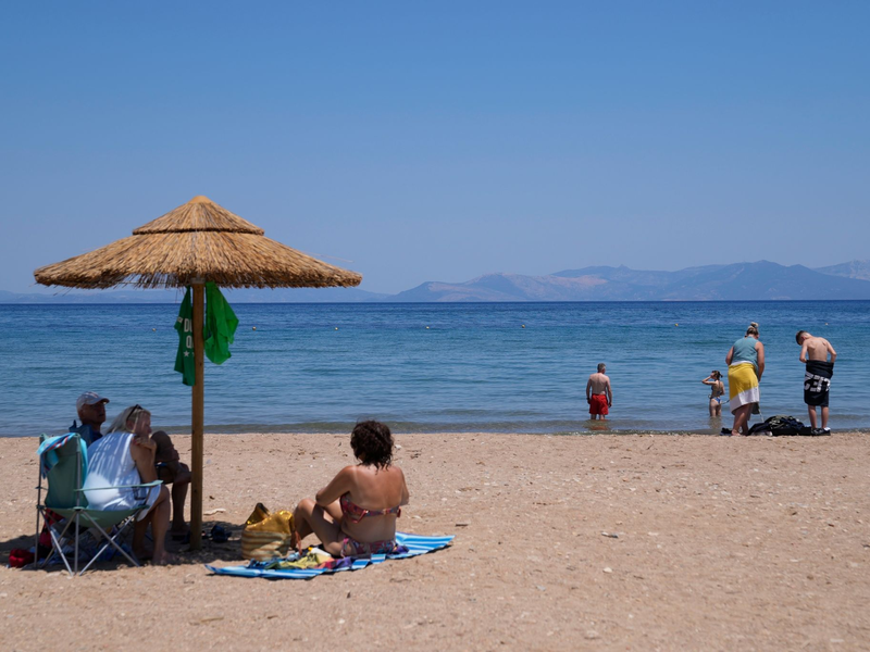 Eine Frau mit einem Handventilator unter einem Sonnenschirm am Strand in Griechenland. Die heißen Mittagsstunden sollten möglichst gemieden werden. - Foto: Thanassis Stavrakis/AP/dpa