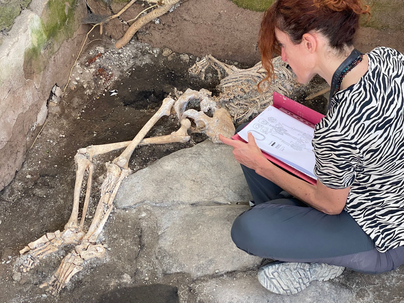 Eine Frau sitzt vor einem Skelett im Archäologiepark Pompeji. - Foto: -/Archäologiepark Pompeji/dpa