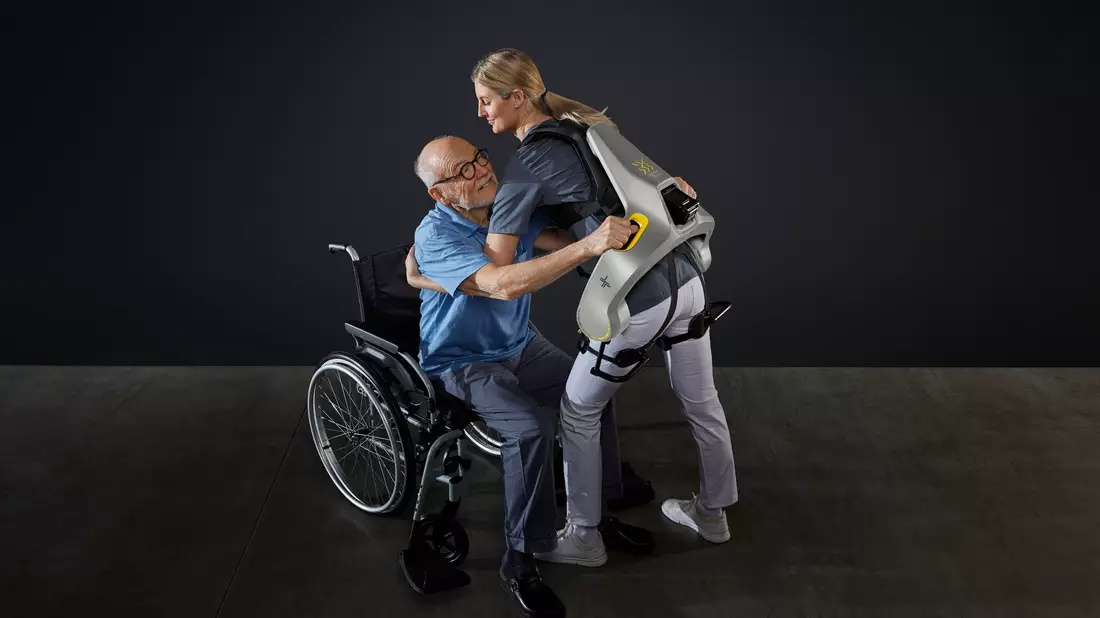 Eine Frau hebt mit Hilfe eines intelligenten Kraftanzugs der Firma German Bionic einen Mann aus einem Rollstuhl. - Bild: German Bionic/dpa