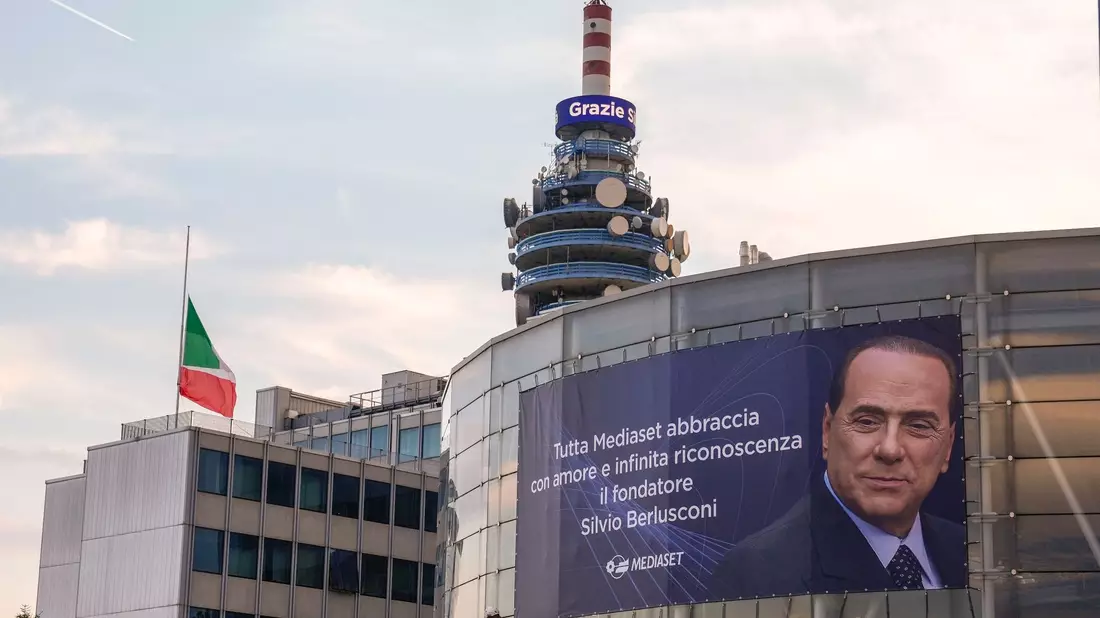 Eine italienische Flagge auf Halbmast weht vor einer Nachricht mit der Aufschrift «Grazie Silvio» auf einem Turm des Fernsehsenders Mediaset neben einem großen Plakat. - Bild: Luca Bruno/AP