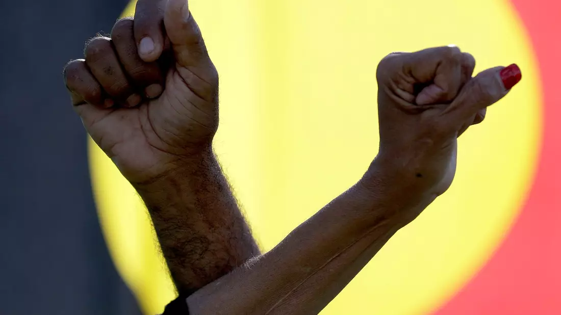 Während einer Kundgebung in Sydney verschränken  zwei Demonstranten vor einer Aborigine-Flagge ihre Arme. - Bild: Rick Rycroft/AP/dpa