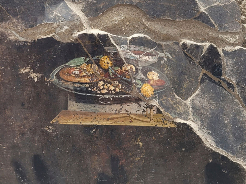 Die Wand eines pompejanischen Hauses mit einem Fresko, das einen Tisch mit Speisen darstellt, darunter auch eine Art Pizza. - Foto: ---/Pompeii Archaelogical Park/AP/dpa