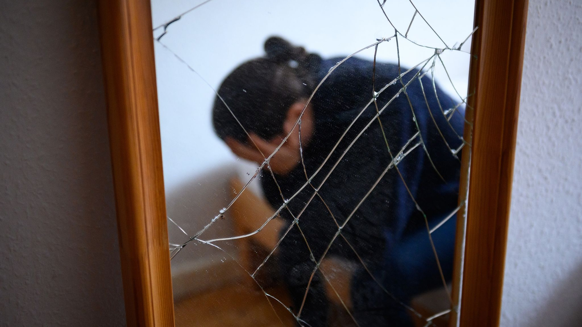 Eine Frau sitzt neben einem gesprungenen Spiegel auf einem Bett und hält ihren Kopf in den Händen. (Symbolbild) - Foto: Jonas Walzberg/dpa