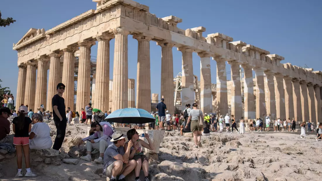 Touristen unter einem Sonnenschirm vor der Akropolis, die inzwischen geschlossen wurde. - Bild: Petros Giannakouris/AP/dpa