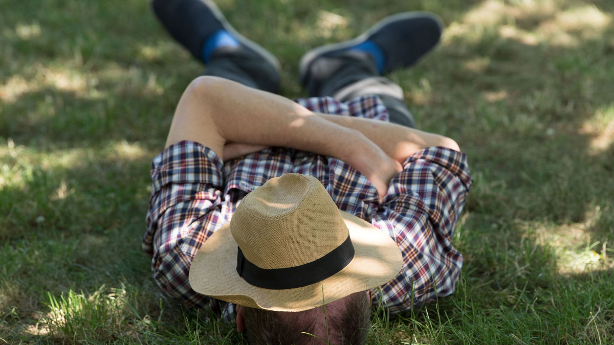 Ein Mann mit einem Sonnenhut auf dem Gesicht liegt auf einer Wiese im Schatten und ruht sich aus. - Foto: Sebastian Kahnert/zb/dpa