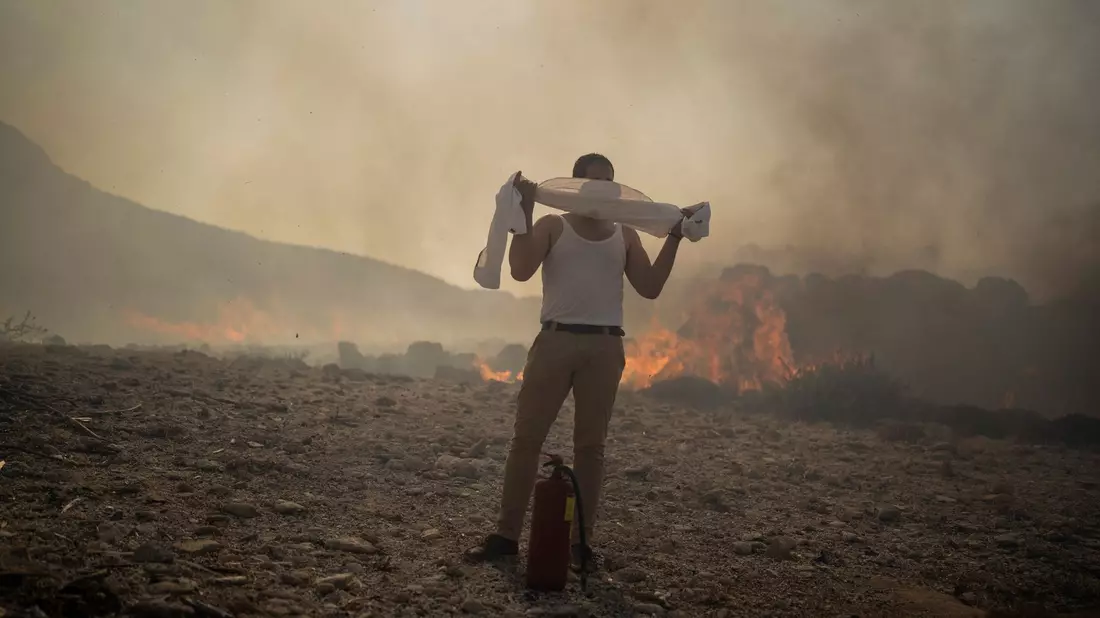 Ein Mann schützt sich mit einem Handtuch vor dem Rauch, während er versucht, ein Feuer nahe des Badeortes Lindos zu löschen. - Bild: Petros Giannakouris/AP/dpa