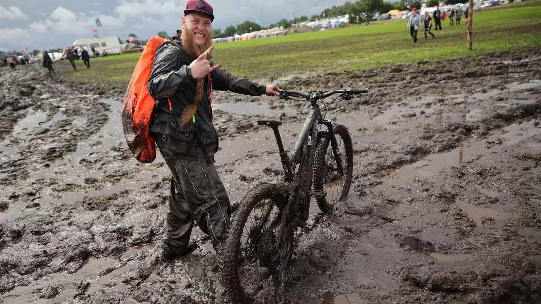 Metal-Fan Lukas Litwin aus Bochum ist mit seinem E-Mountainbike auf dem schlammigen Festivalgelände unterwegs. Noch ist unklar, wie viele der erwarteten 85.000 Metalfans tatsächlich auf Wackens Ackern feiern. - Bild: Christian Charisius/dpa
