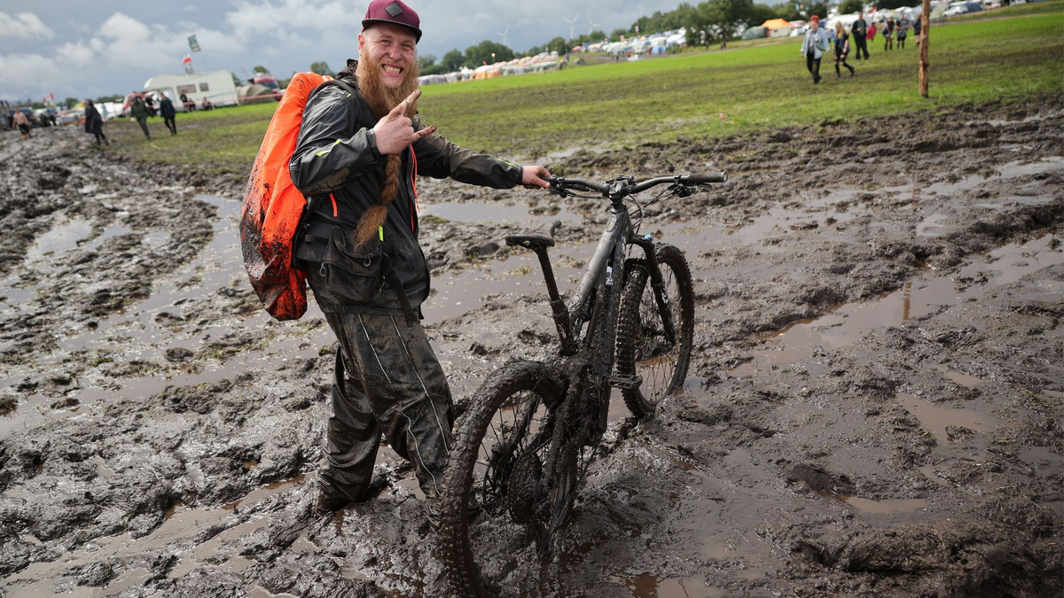 Metal-Fan Lukas Litwin aus Bochum ist mit seinem E-Mountainbike auf dem schlammigen Festivalgelände unterwegs. Noch ist unklar, wie viele der erwarteten 85.000 Metalfans tatsächlich auf Wackens Ackern feiern. - Foto: Christian Charisius/dpa