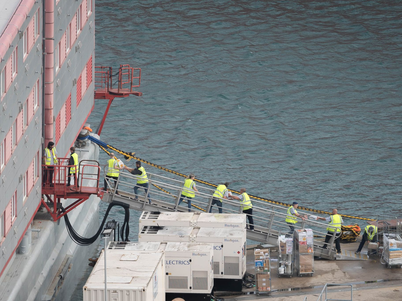Ein Schleppschiff passiert im Hafen von Portland in Dorset das Schiff «Bibby Stockholm», das bis zu 500 Menschen aufnehmen soll. - Foto: James Manning/PA Wire/dpa