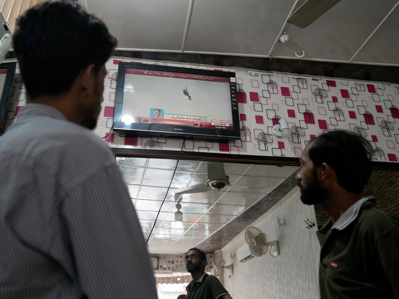Menschen in einem Friseur-Salon verfolgen eine Nachrichtensendung, in der über eine riskante Rettungsmission an der Seilbahn berichtet wird. - Foto: K.M. Chaudary/AP/dpa