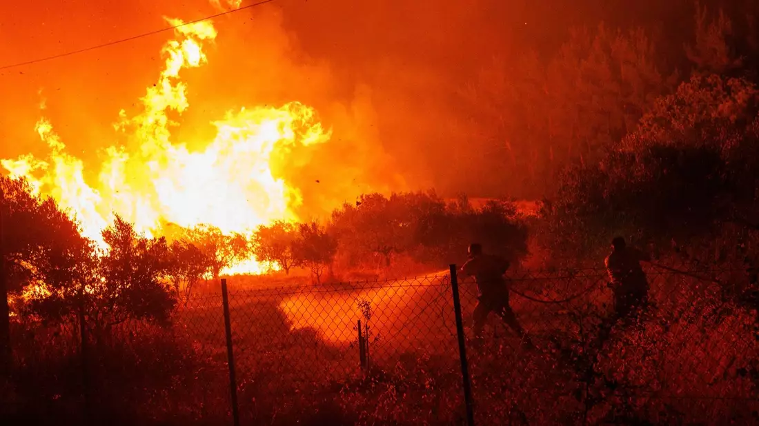 Ein Hubschrauber wirft Wasser über dem Dorf Avantas ab. Stürmische Winde fachen die Flammen der Waldbrände in ganz Griechenland an. - Bild: Achilleas Chiras/AP