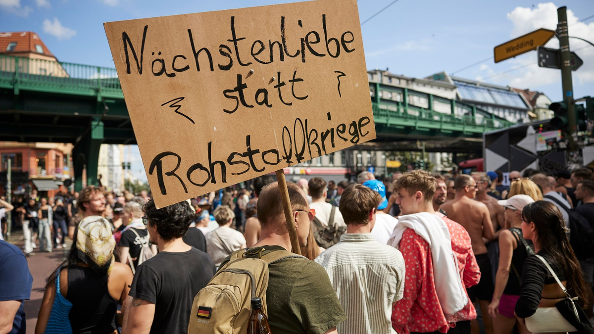 Ein Feiernder trägt ein Schild mit der Aufschrift «Nächstenliebe statt Rohstoffkriege» bei der Technoparade Zug der Liebe durch Berlin. - Foto: Joerg Carstensen/dpa