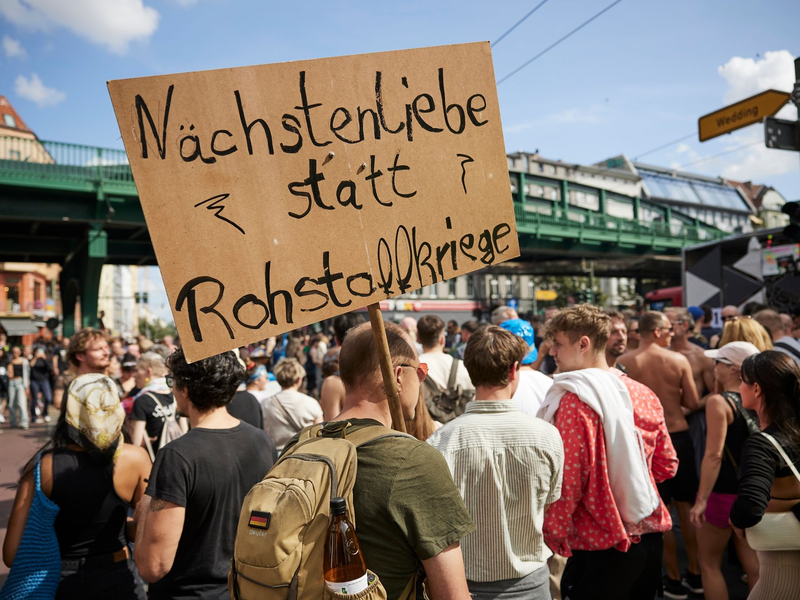 Ein Feiernder trägt ein Schild mit der Aufschrift «Nächstenliebe statt Rohstoffkriege» bei der Technoparade Zug der Liebe durch Berlin. - Foto: Joerg Carstensen/dpa