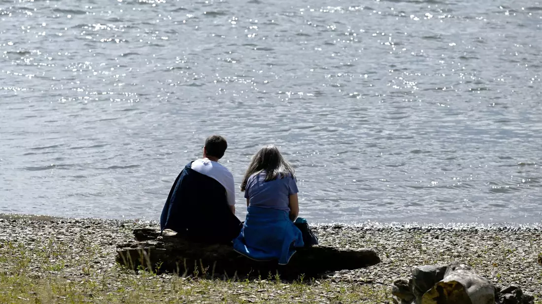 Spaziergänger sitzen am Ufer des Rheins und genießen die Sonne. - Bild: Roberto Pfeil/dpa