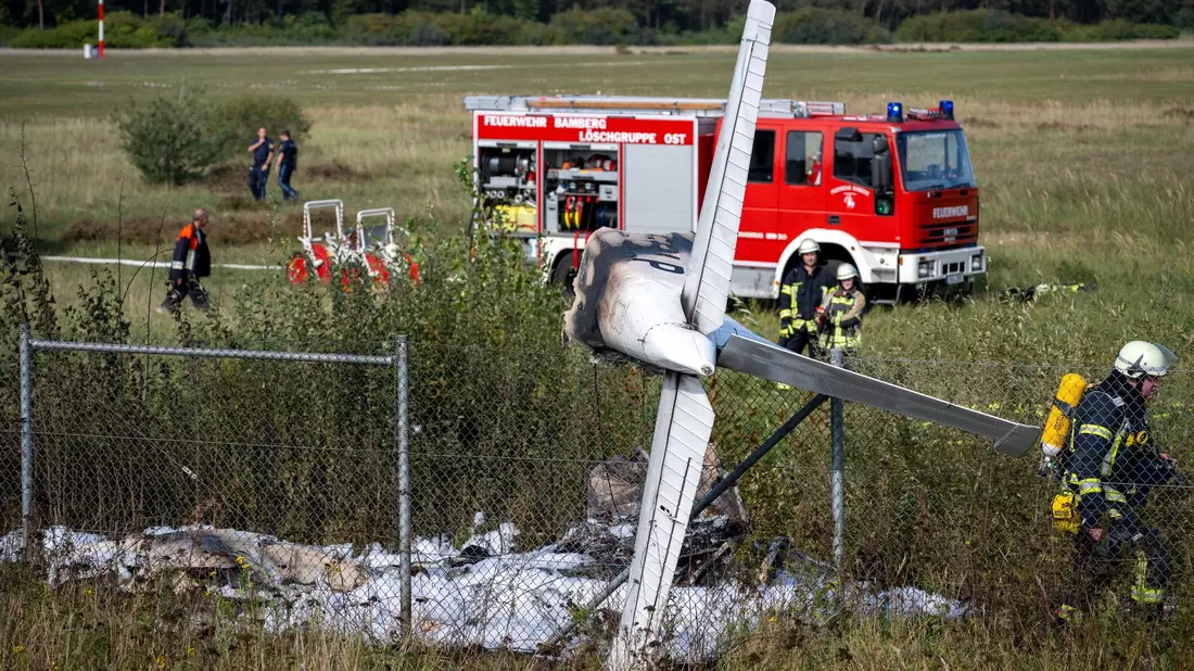 Trümmerteile eines Flugzeugs hängen in einem Zaun, während Einsatzkräfte der Feuerwehr an der Absturzstelle stehen. - Bild: Pia Bayer/dpa