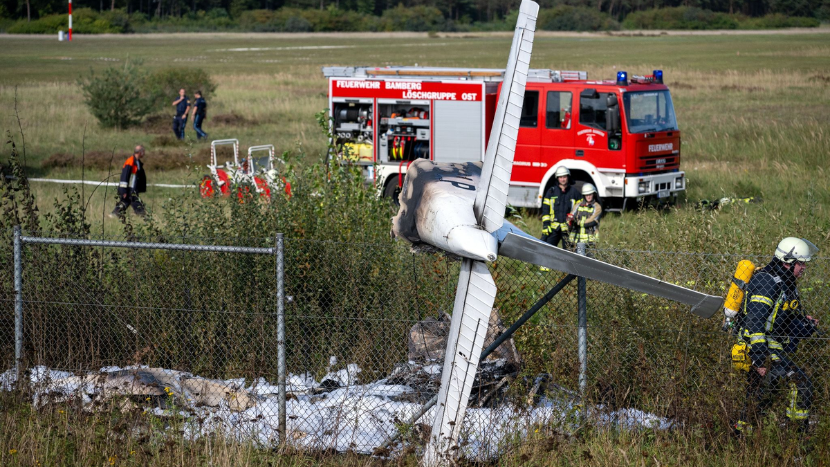 Trümmerteile eines Flugzeugs hängen in einem Zaun, während Einsatzkräfte der Feuerwehr an der Absturzstelle stehen. - Foto: Pia Bayer/dpa