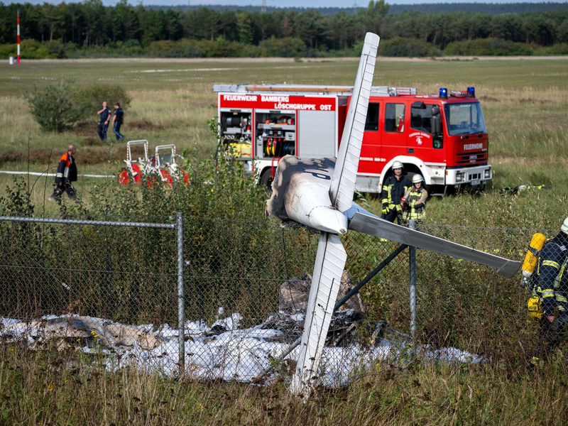 Trümmerteile eines Flugzeugs hängen in einem Zaun, während Einsatzkräfte der Feuerwehr an der Absturzstelle stehen. - Foto: Pia Bayer/dpa