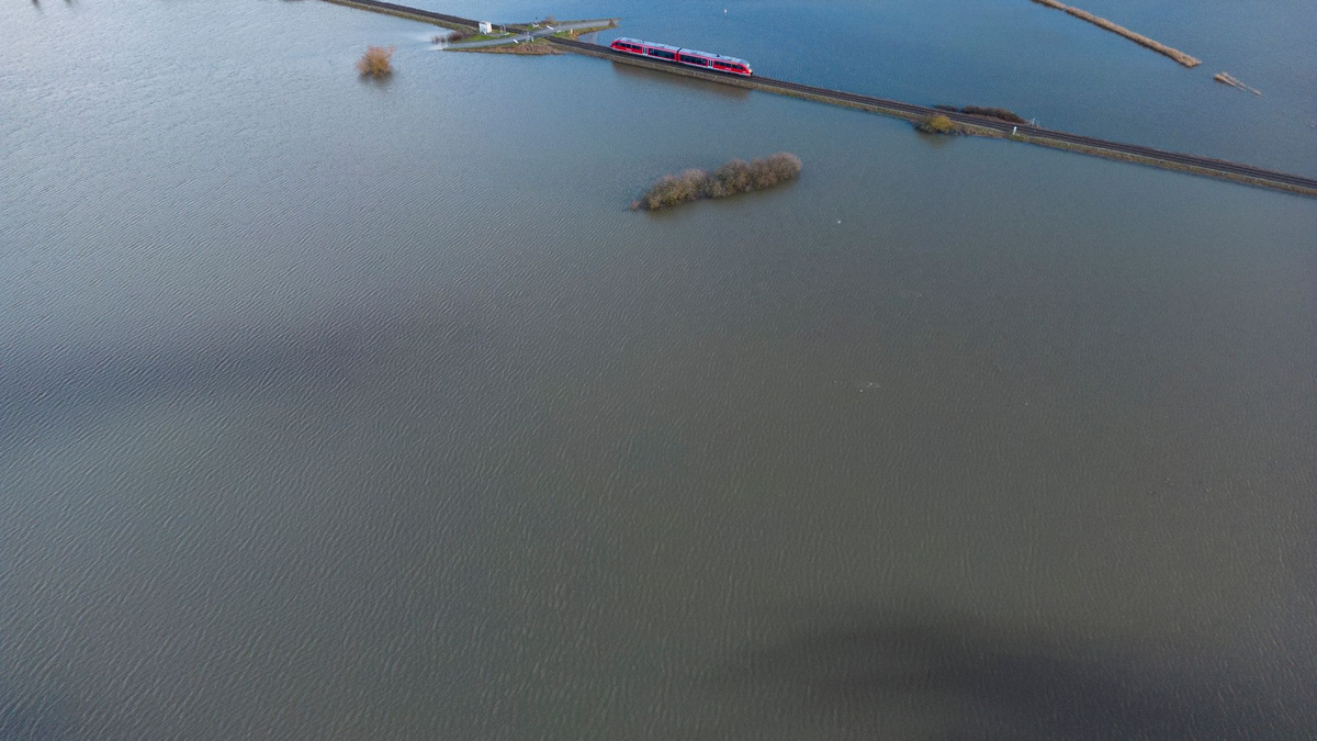 Ein Personenzug der Deutschen Bahn fährt im Januar 2023 durch eine überschwemmte Flussniederung bei Eichen in Hessen. - Foto: Boris Roessler/dpa