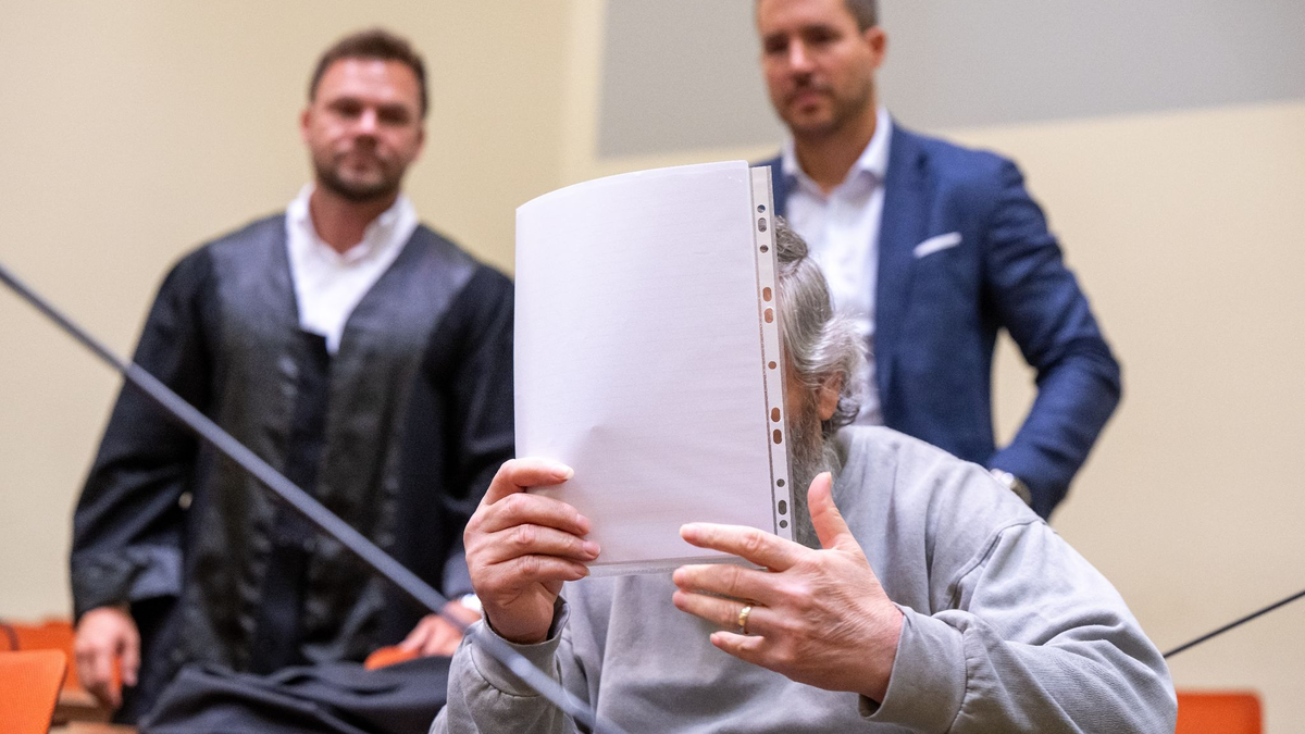 Der Angeklagte hält sich im Gerichtssaal ein Blatt Papier vor das Gesicht. Im Hintergrund stehen seine Anwälte. - Foto: Peter Kneffel/dpa