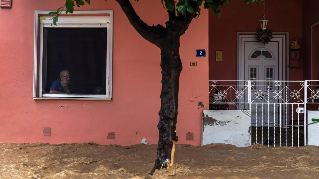 Ein Anwohner blickt aus dem Fenster seines Hauses auf die überschwemmte Straße. - Bild: Petros Giannakouris/AP/dpa