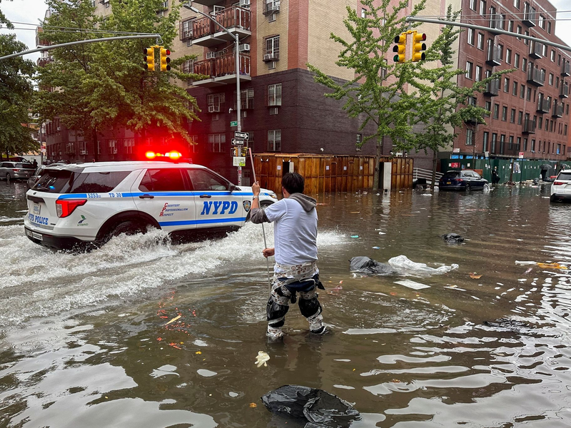 Hochwasser auf den Straßen von New York: Im Stadtteil Brooklyn versucht ein Mann, mit einem  Stock einen Abfluss zu reinigen, während ein Polizeiwagen vorbeifährt. - Foto: Jake Offenhartz/AP/dpa