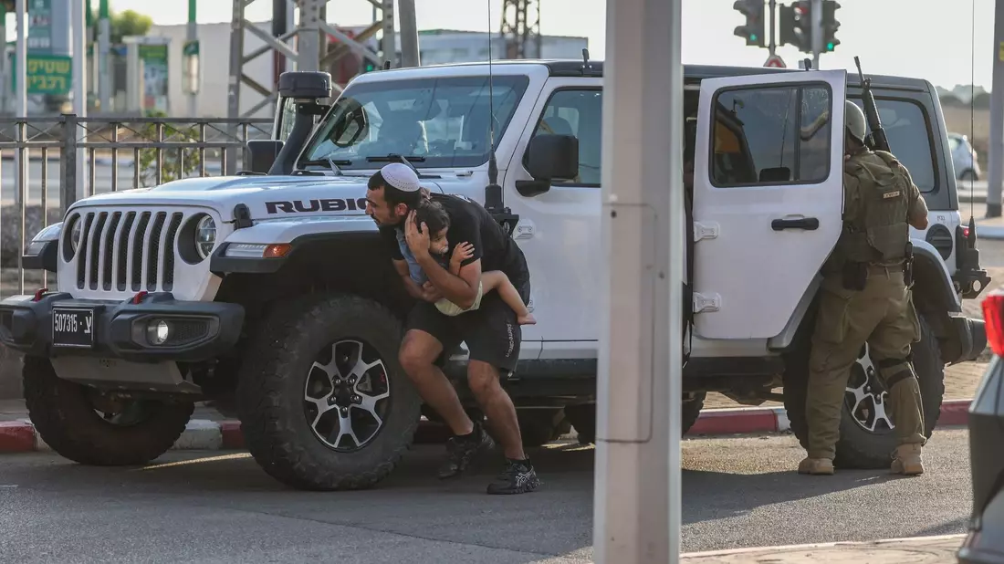 Ein Mann hält in Ashkelon ein Kind im Arm und sucht bei einem neuen Raketenangriff aus Gaza hinter einem Fahrzeug Deckung. - Bild: Ilia yefimovich/dpa