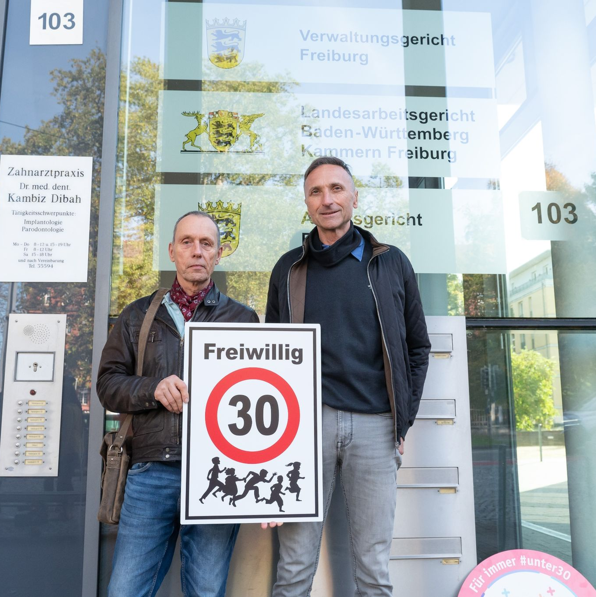 Christian Kronbitter (l) und Erich Maier vor dem Verwaltungsgericht in Freiburg mit einem «Freiwillig 30»-Schild. - Foto: Silas Stein/dpa