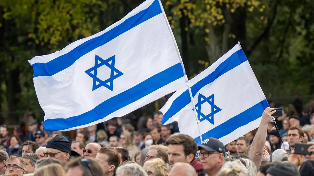 Teilnehmer der Kundgebung „Aufstehen gegen Terror, Hass und Antisemitismus – in Solidarität und Mitgefühl mit Israel“ stehen umhüllt in eine israelische Flagge vor dem Brandenburger Tor in Berlin. - Bild: Monika Skolimowska/dpa