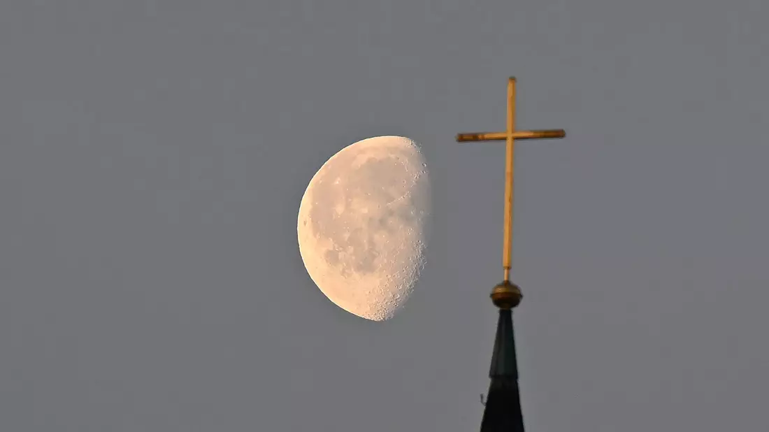Der Mond im Morgenlicht hinter einem Kreuz auf einem Kirchturm. - Bild: Bernd Weißbrod/dpa