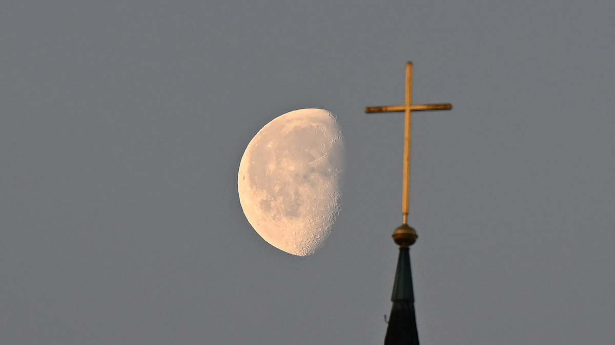 Der Mond im Morgenlicht hinter einem Kreuz auf einem Kirchturm. - Foto: Bernd Weißbrod/dpa