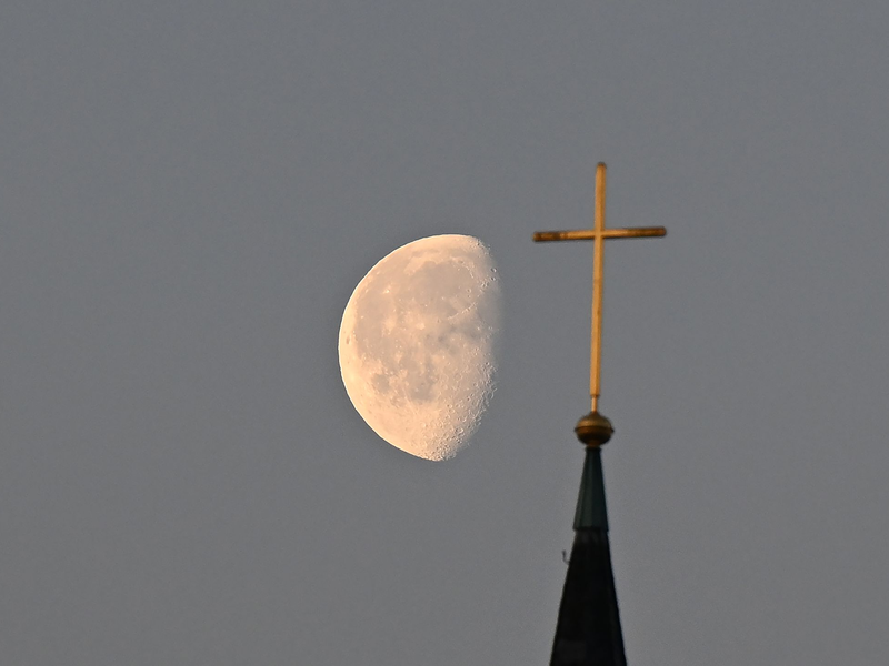 Der Mond im Morgenlicht hinter einem Kreuz auf einem Kirchturm. - Foto: Bernd Weißbrod/dpa