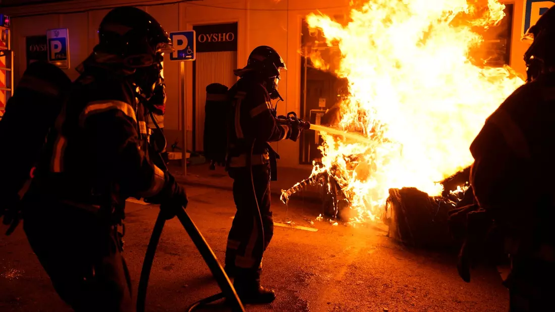 Feuerwehrleute müssen einen von Demonstranten angezündeten Müllcontainer löschen. - Bild: Manu Fernandez/AP/dpa