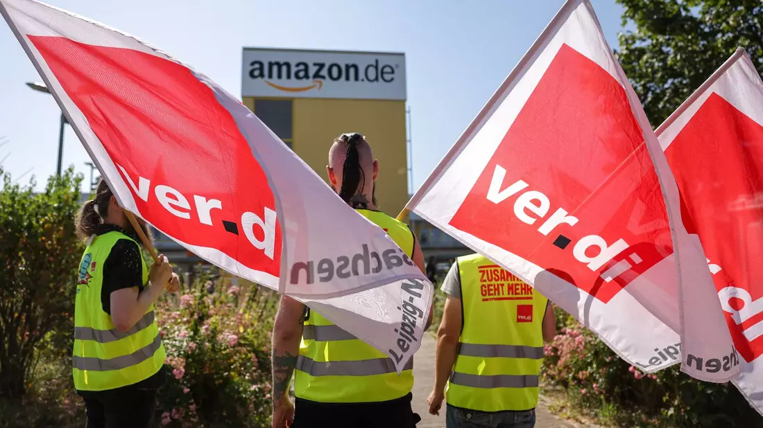 Streikende stehen mit Verdi-Flaggen vor einem Verteilzentrum des Online-Händlers Amazon in Leipzig (Archivbild). - Bild: Jan Woitas/dpa