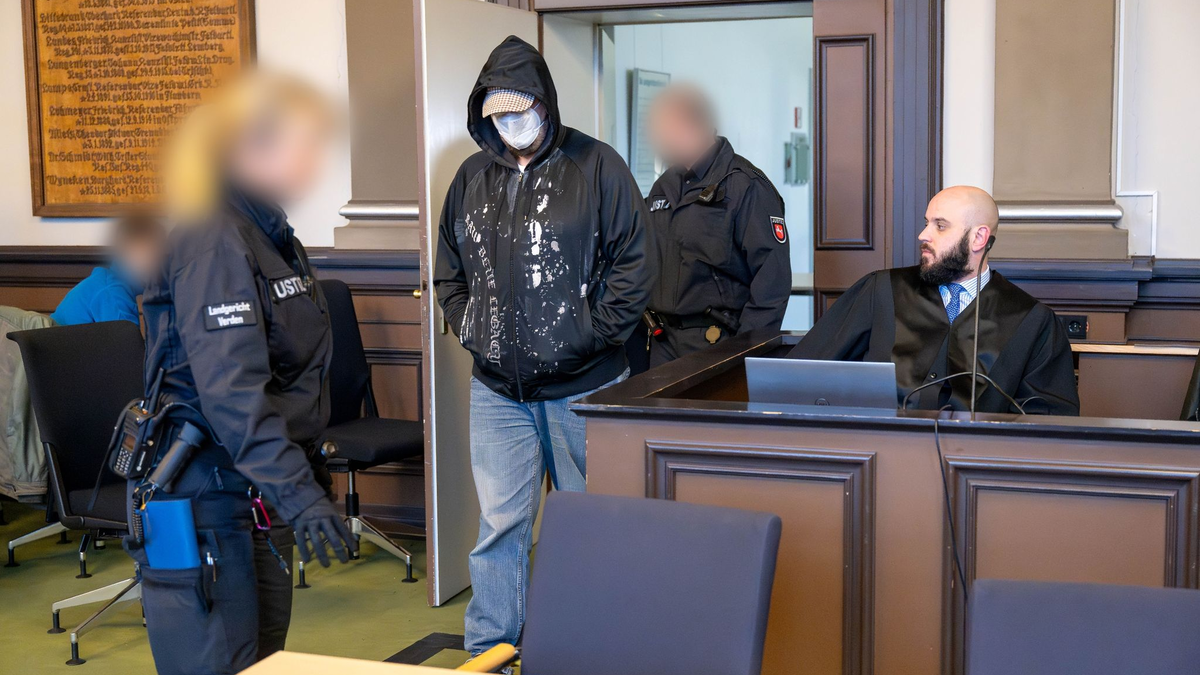 Einer der drei Angeklagten (M) wird vor Prozessbeginn von Justizbeamten in den Gerichtssaal geführt. - Foto: Sina Schuldt/dpa