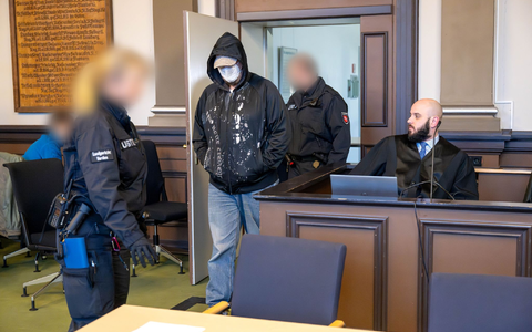 Einer der drei Angeklagten (M) wird vor Prozessbeginn von Justizbeamten in den Gerichtssaal geführt. - Foto: Sina Schuldt/dpa