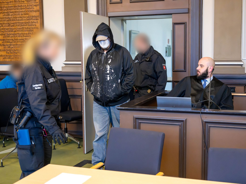 Einer der drei Angeklagten (M) wird vor Prozessbeginn von Justizbeamten in den Gerichtssaal geführt. - Foto: Sina Schuldt/dpa