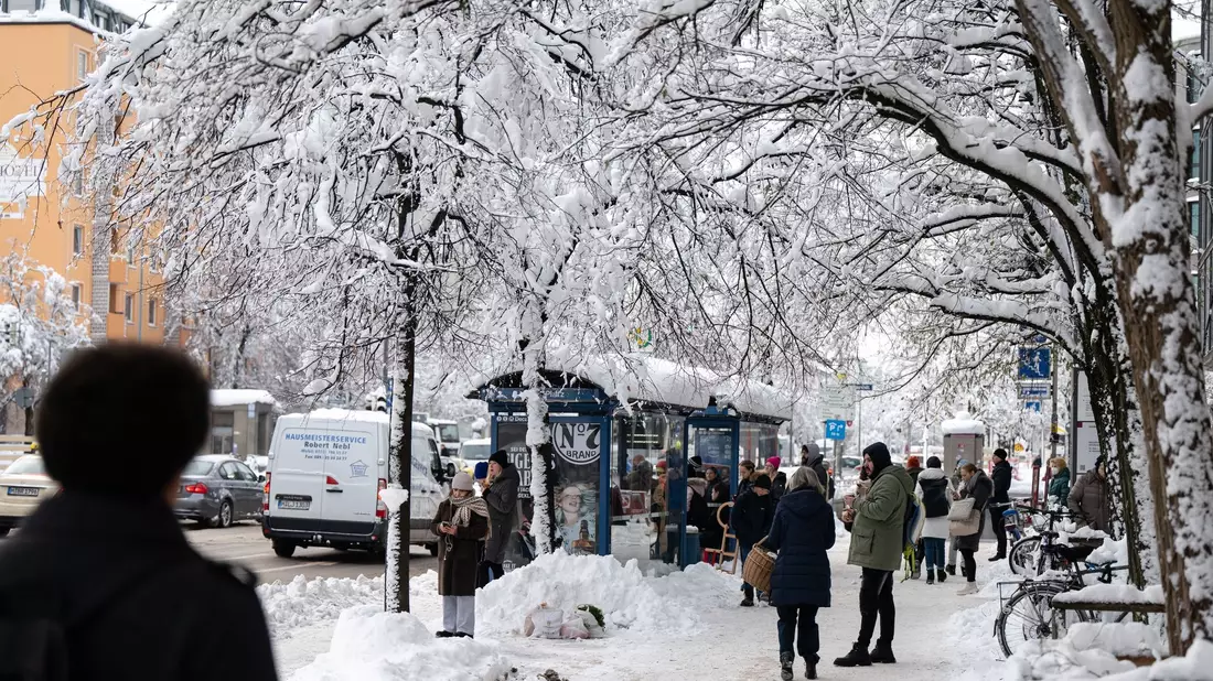 Fahrgäste warten an einer verschneiten Haltestelle auf einen Bus. - Bild: Lukas Barth/dpa
