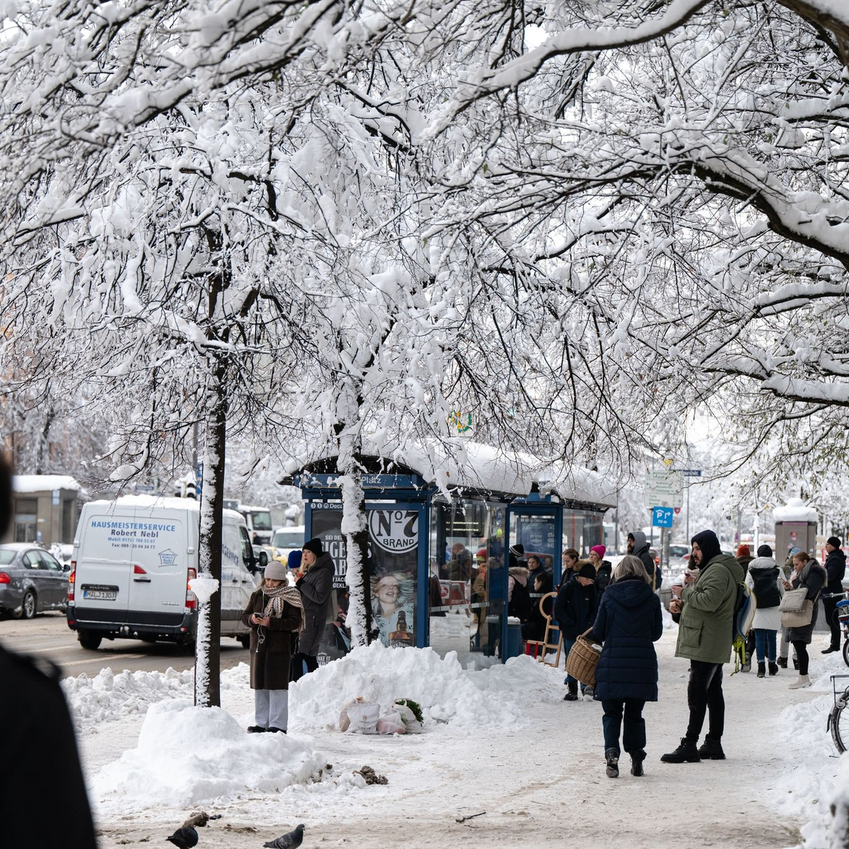 Fahrgäste warten an einer verschneiten Haltestelle auf einen Bus. - Foto: Lukas Barth/dpa