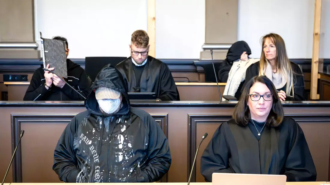 Die drei Angeklagten (l, vorne l sowie hinten r) sitzen vor Prozessbeginn mit ihren Anwältinnen und Anwälten im Gerichtssaal. - Bild: Sina Schuldt/dpa