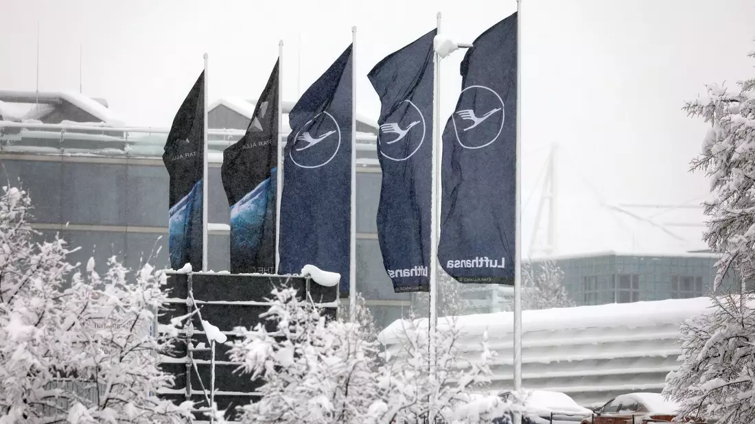 Fahnen der Lufthansa wehen am  Flughafen München im Schneetreiben. Die deutsche Fluggesellschaft kauft neue Maschinen ein. - Bild: Karl-Josef Hildenbrand/dpa