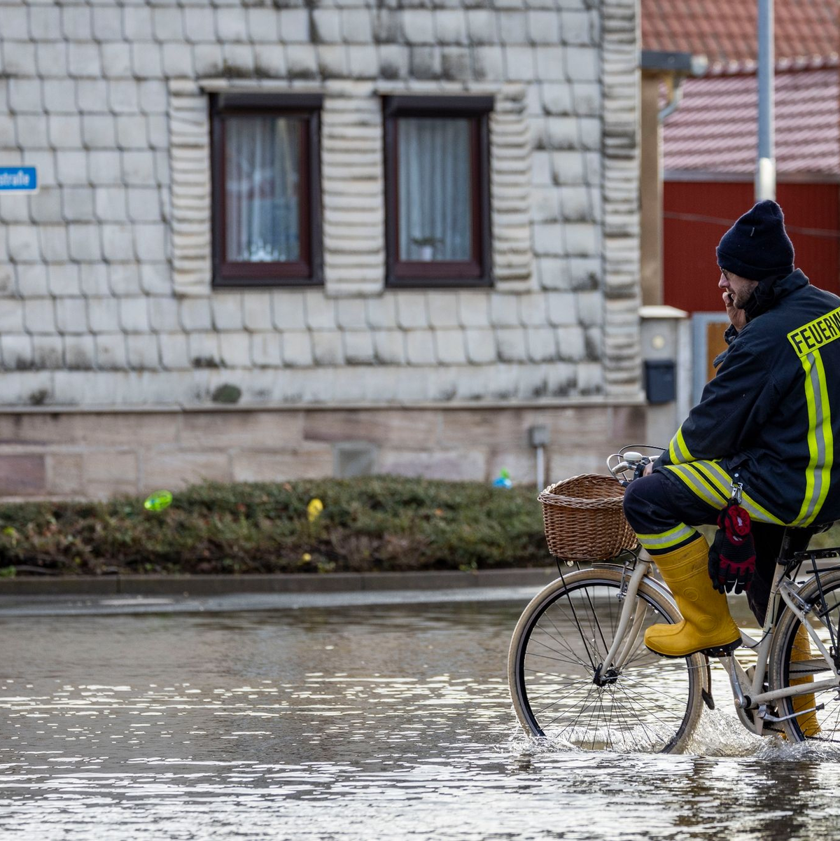 Ein Feuerwehrmann auf einem Fahrrad in einer überschwemmten Straße in Windehausen. - Foto: Christoph Reichwein/dpa