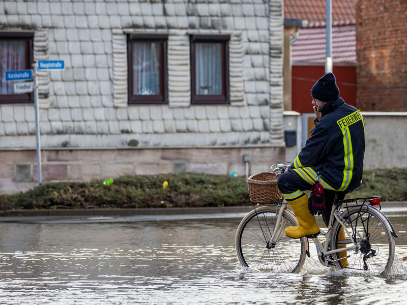 Ein Feuerwehrmann auf einem Fahrrad in einer überschwemmten Straße in Windehausen. - Foto: Christoph Reichwein/dpa