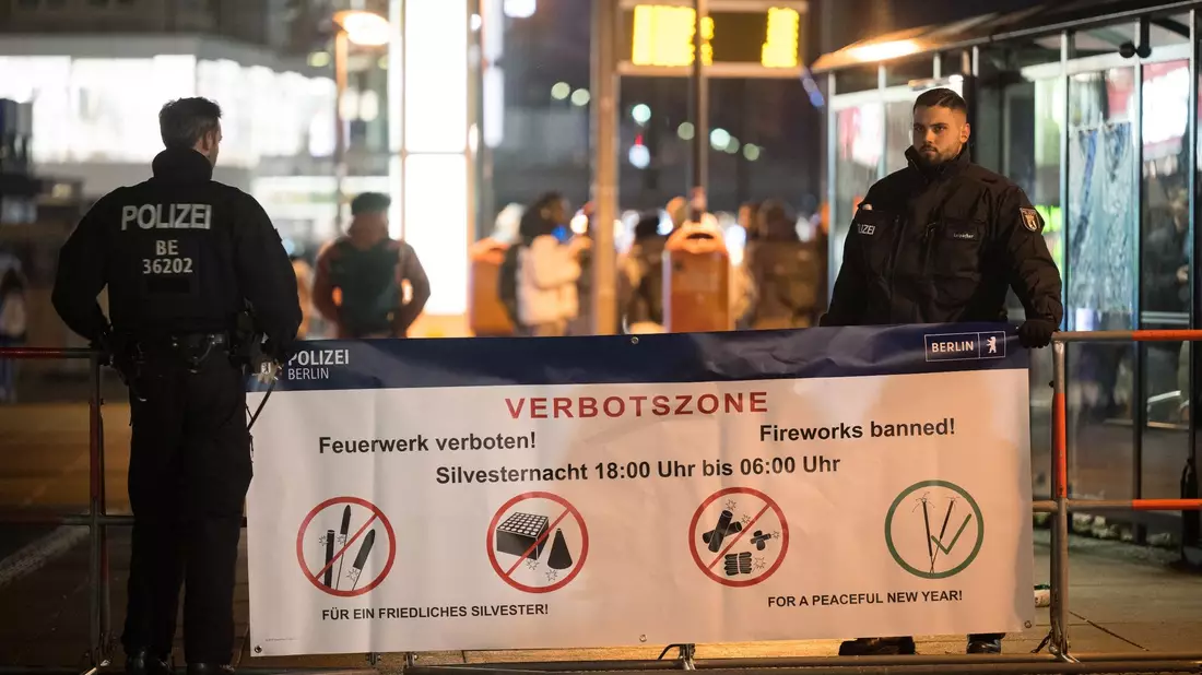 Polizeibeamte stehen an einem Plakat, das auf die Böllerverbotszone am Berliner Alexanderplatz hinweist. - Bild: Sebastian Christoph Gollnow/dpa