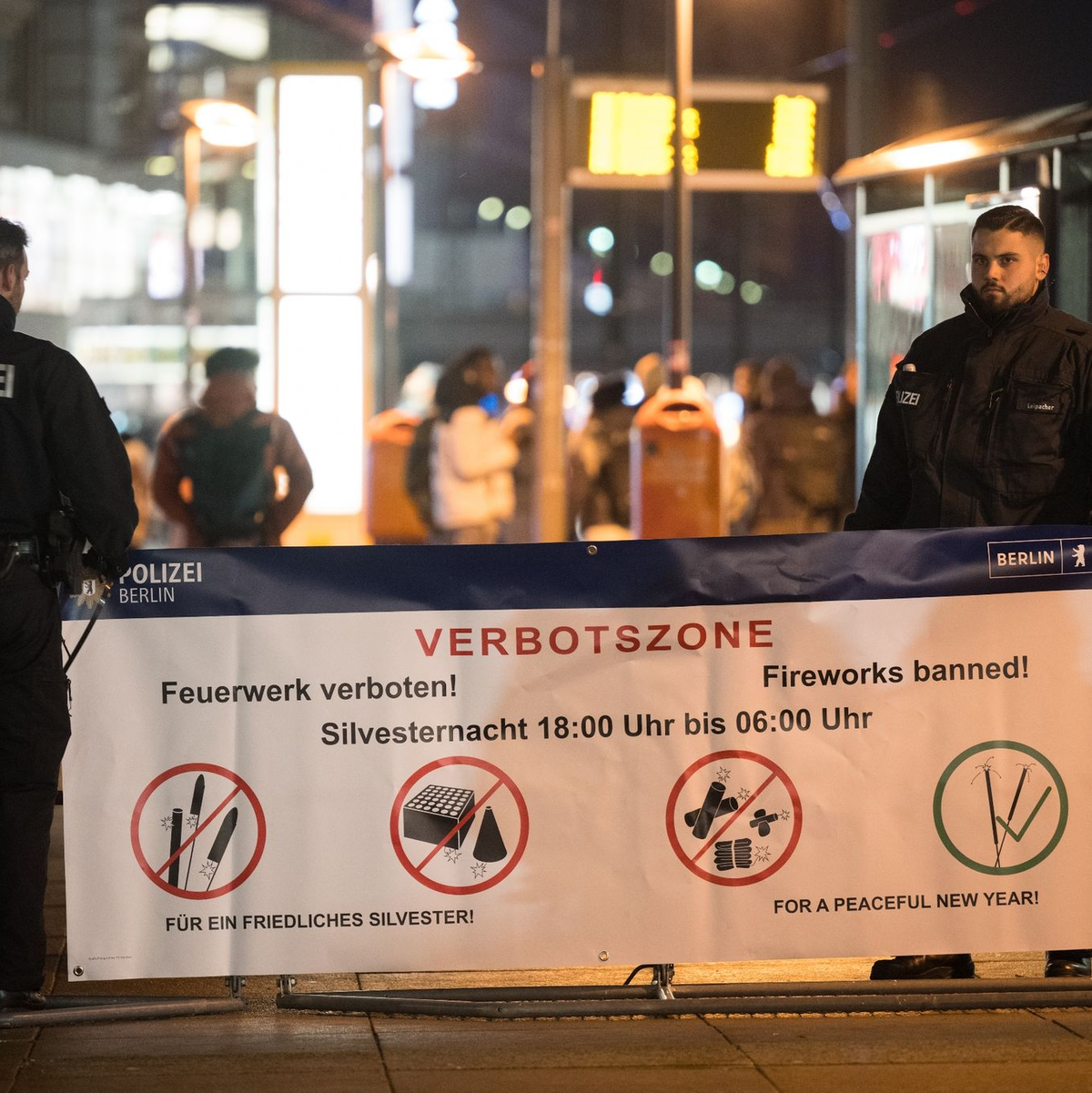 Polizeibeamte stehen an einem Plakat, das auf die Böllerverbotszone am Berliner Alexanderplatz hinweist. - Foto: Sebastian Christoph Gollnow/dpa