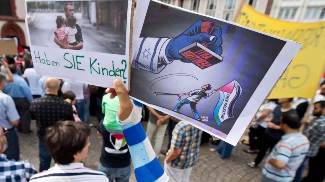 Ein Junge hält ein Plakat hoch, auf dem ein propalästinensischer Demonstrant unter einem Stempel mit der Aufschrift  Anti Semitismus steht. - Bild: picture alliance / dpa