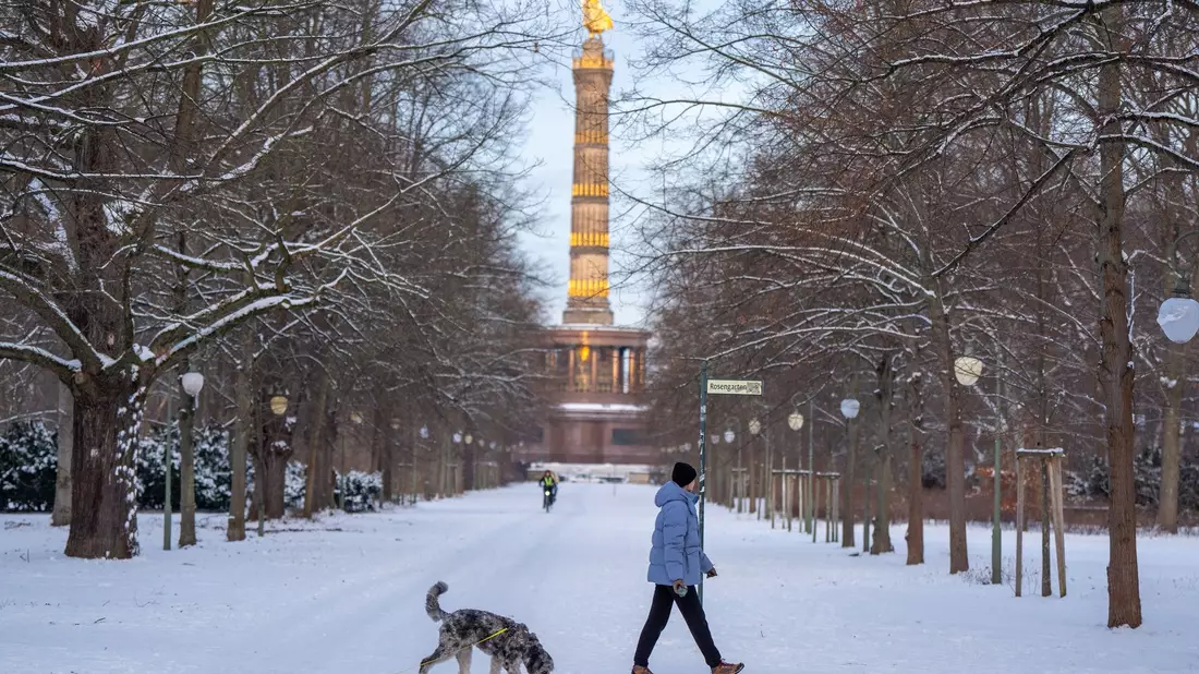 Eine Frau geht mit ihrem Hund vor der Kulisse der Siegessäule im verschneiten Tiergarten in Berlin spazieren. - Bild: Soeren Stache/dpa