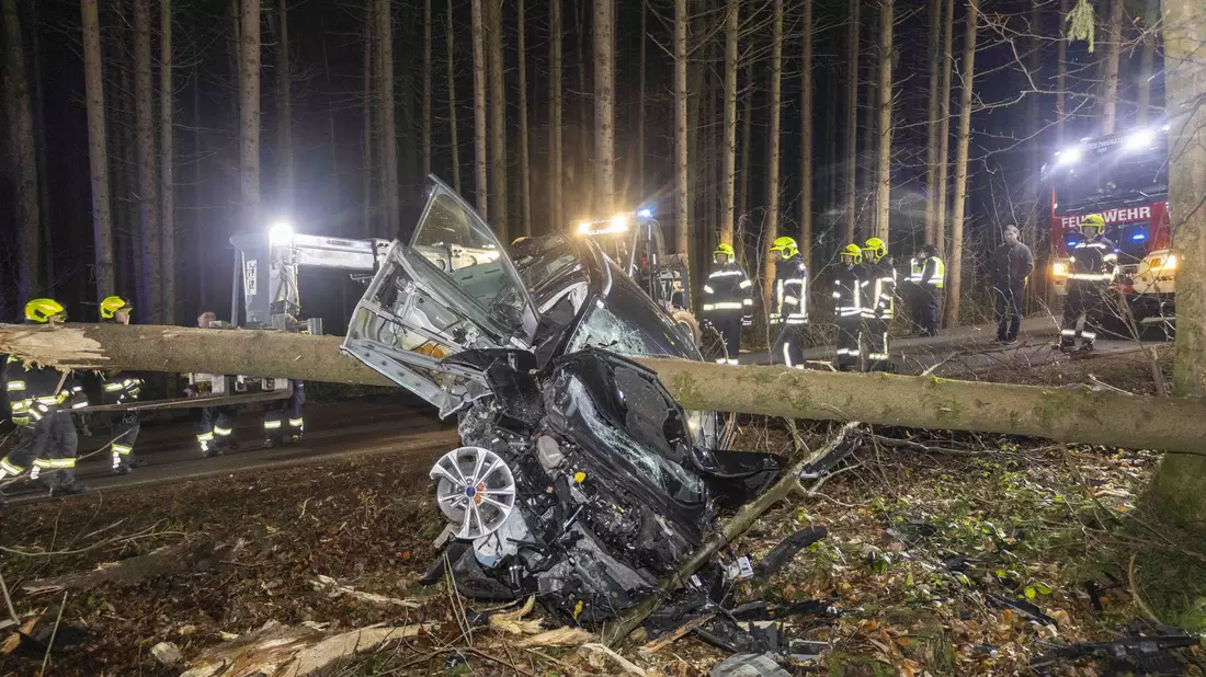 Feuerwehrleute betrachten das Auto im österreichischen Aurolzmünster, das in einen liegenden Baumstamm krachte. - Bild: Daniel Scharinger/APA/dpa